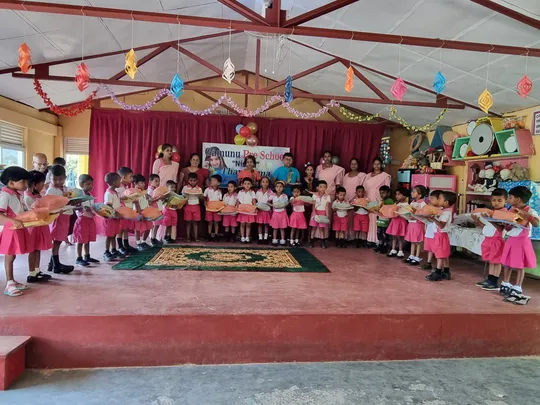 Schoolchildren standing in a semicircle, holding gifts they have received as donations. Adults are standing behind the semicircle.