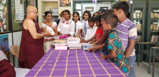 Children standing around a table, as well as a Buddhist monk. The table has a purple tablecloth with a yellow checked pattern, and has stacks of books on it.