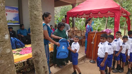 Children queueing up. At the front of the queue, a child is receiving a blue backpack from two adult women who are smiling. In the background, another woman is standing at a podium with a microphone.