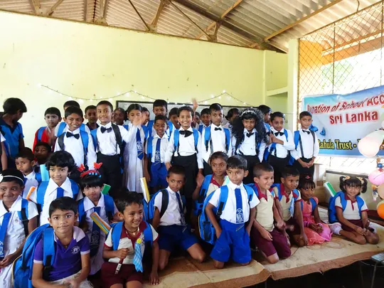 A group of schoolchildren smiling and waving at the camera. Each child is wearing a blue backpack.